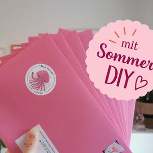 Könnte beinhalten: Ein Stapel hellrosa Umschläge mit einem runden Aufkleber, der eine rosa Qualle und den Text Marli Design Creative Mail Club zeigt. Eine rosa, gewellte Grafiküberlagerung enthält den Text mit Sommer DIY und ein Herzsymbol.