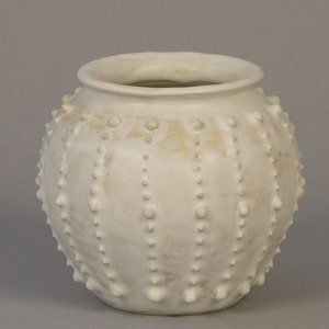 Peut inclure: Vase en céramique de couleur crème, avec un corps rond et une large ouverture. La surface est texturée avec des rangées de petites bosses arrondies et espacées régulièrement. Le vase a un aspect légèrement vieilli.