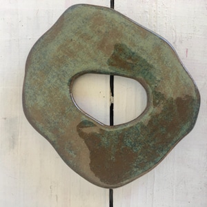 Può includere: Un pezzo di ceramica fatto a mano, di forma organica irregolare, con un'apertura centrale. La ceramica presenta una tavolozza di colori terrosi e screziati di verdi e marroni, con una finitura lucida. Il design suggerisce un oggetto d'arte decorativo o funzionale.