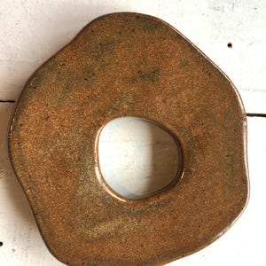 Könnte beinhalten: Ein handgefertigter, organisch geformter Keramikteller mit einem zentralen Loch. Der Teller hat eine gesprenkelte, erdfarbene braune Glasur mit Goldtönen und ein glänzendes Finish. Das Design erinnert an einen Donut oder einen großen Ring.