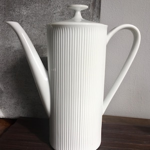 Op de afbeelding: Witte keramische koffiekan met een geribbeld ontwerp en een deksel. De kan heeft een tuit en een handvat.