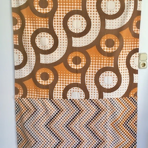 Könnte beinhalten: Ein Vintage-Vorhang mit Retro-Design. Der obere Teil zeigt große braune Kreise und orangefarbene Punkte auf weißem Hintergrund. Der untere Teil hat ein braunes, orangefarbenes und weißes Zickzackmuster. Der Vorhang hängt an einer weißen Wand.