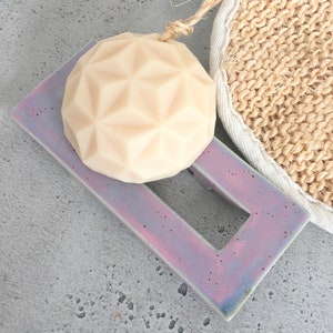 Puede incluir: Un jabón beige claro con un patrón geométrico descansa sobre un jabonera rectangular iridiscente con un centro recortado. También se ve una esponja de luffa natural con un borde blanco y una cuerda de yute.