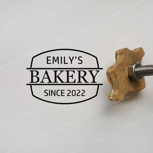 Puede incluir: Un logotipo de Emily's Bakery, fundada en 2022, se muestra sobre un fondo blanco. El logotipo negro presenta el nombre de la panadería y el año de fundación. A la derecha, un objeto metálico dorado con un tubo plateado flexible.