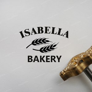 May include: A gold metal stamp with a black imprint of the text "Isabella Bakery" and a wheat stalk design.