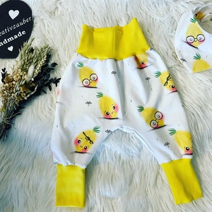 Könnte beinhalten: Weißes Babyhosen- und Lätzchen-Set mit einem sich wiederholenden Muster aus lächelnden gelben Ananas mit Brille. Die Hose hat gelbe Bündchen und einen gelben Bund. Das Lätzchen hat ein passendes Ananas-Design. Der Text "T.S. - Kreativzauber Handmade" steht auf einem herzförmigen Schild.