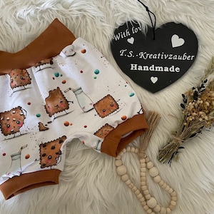 Könnte beinhalten: Weiße Baby-Shorts mit braunem Bund. Die Shorts haben einen Aufdruck von Comic-Keksen und Milch. Der Text "T.S. - Kreativzauber Handmade" befindet sich auf einem schwarzen herzförmigen Schild.