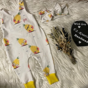 Könnte beinhalten: Weißer Baby-Strampler und passendes Stirnband-Set. Der Strampler hat ein sich wiederholendes Muster aus Cartoon-Ananas mit Brille und Schleifen. Die Bündchen sind gelb. Ein schwarzes herzförmiges Etikett trägt die Aufschrift "With love T.S.-Kreativzauber Handmade".