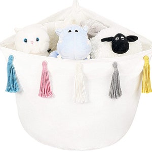 Puede incluir: Cesta de almacenamiento de lona blanca con cinco borlas de colores. La cesta está llena de animales de peluche, incluyendo un gato blanco, un hipopótamo azul y una oveja blanca con la cara negra.