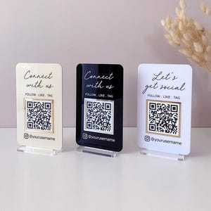 Può includere: Tre supporti in acrilico per codici QR di social media in crema, nero e bianco. Ogni supporto ha un codice QR, il testo "Connect with us" o "Let's get social" e il nome utente "@yourusername".