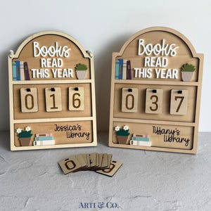 May include: Two arched wooden wall plaques with the text Books READ THIS YEAR. Each features three hanging wooden number tiles, a potted plant, and a stack of books. One plaque reads Jessica's library, the other Tiffany's library. Extra number tiles lie below.