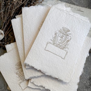 Puede incluir: Una pila de cinco tarjetas de papel en blanco, de color blanquecino y texturado, con un escudo decorativo en relieve de color dorado en el centro de la tarjeta superior. El escudo presenta un dise&ntilde;o floral y un escudo con un le&oacute;n.