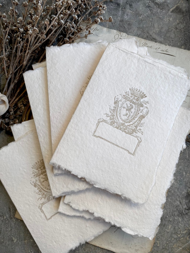 Puede incluir: Una pila de cinco tarjetas de papel en blanco, de color blanquecino y texturado, con un escudo decorativo en relieve de color dorado en el centro de la tarjeta superior. El escudo presenta un dise&ntilde;o floral y un escudo con un le&oacute;n.