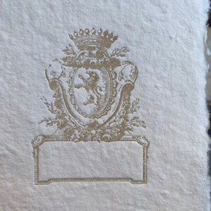 Puede incluir: Una tarjeta de papel blanco, de estilo vintage, en blanco, con un escudo decorativo en relieve de color dorado que representa un le&oacute;n y una corona.