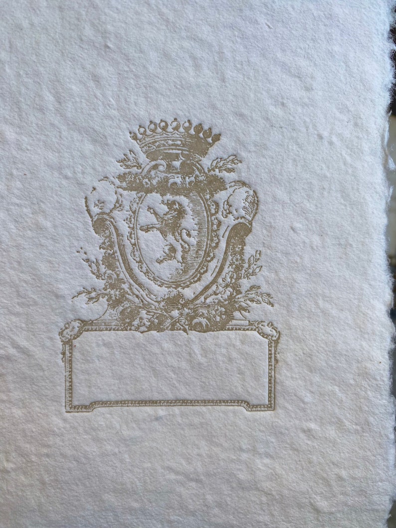 Puede incluir: Una tarjeta de papel blanco, de estilo vintage, en blanco, con un escudo decorativo en relieve de color dorado que representa un le&oacute;n y una corona.