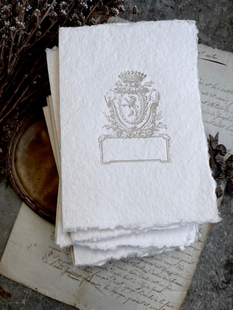 Puede incluir: Una pila de papel blanco texturizado con un escudo decorativo en relieve que presenta un le&oacute;n y una corona. El escudo est&aacute; rodeado de un dise&ntilde;o floral y un espacio rectangular en blanco.