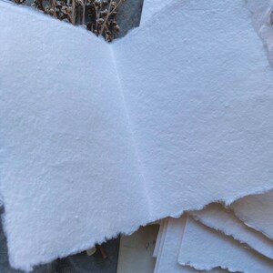 Puede incluir: Una pila de papel blanco hecho a mano con bordes &aacute;speros. El papel es texturizado y tiene un color ligeramente blanquecino. El papel est&aacute; dispuesto en una pila con la hoja superior doblada por la mitad.