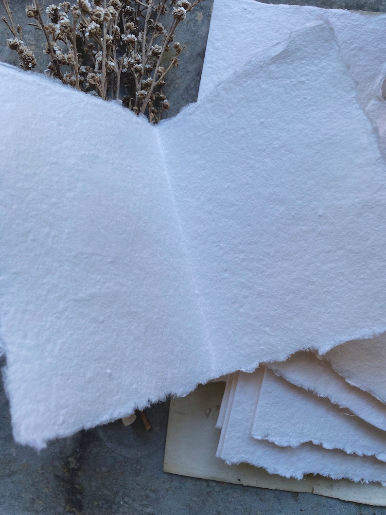 Puede incluir: Una pila de papel blanco hecho a mano con bordes &aacute;speros. El papel es texturizado y tiene un color ligeramente blanquecino. El papel est&aacute; dispuesto en una pila con la hoja superior doblada por la mitad.