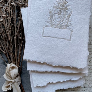 Puede incluir: Pila de cuatro hojas de papel blanco texturizado, hechas a mano, con un escudo gris decorativo en relieve que presenta una corona y un escudo. El escudo tiene un espacio rectangular en blanco para escribir.