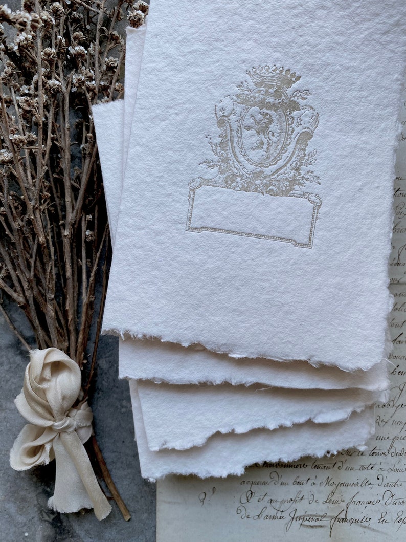 Puede incluir: Pila de cuatro hojas de papel blanco texturizado, hechas a mano, con un escudo gris decorativo en relieve que presenta una corona y un escudo. El escudo tiene un espacio rectangular en blanco para escribir.