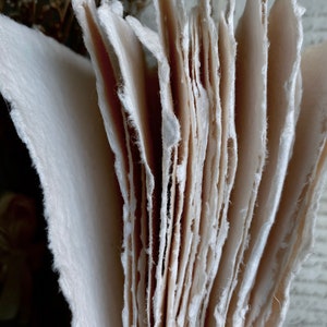Puede incluir: Una pila de hojas de papel hechas a mano con bordes &aacute;speros e irregulares. El papel es de color blanco crema y tiene una superficie texturizada.