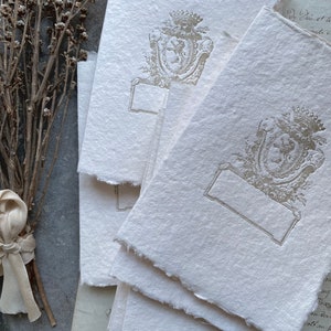 Puede incluir: Una pila de tarjetas de papel en blanco, de color blanquecino, con textura, con un escudo decorativo en relieve plateado que presenta una corona y un le&oacute;n. El escudo est&aacute; rodeado de un dise&ntilde;o floral. Las tarjetas tienen un borde irregular y desgarrado.