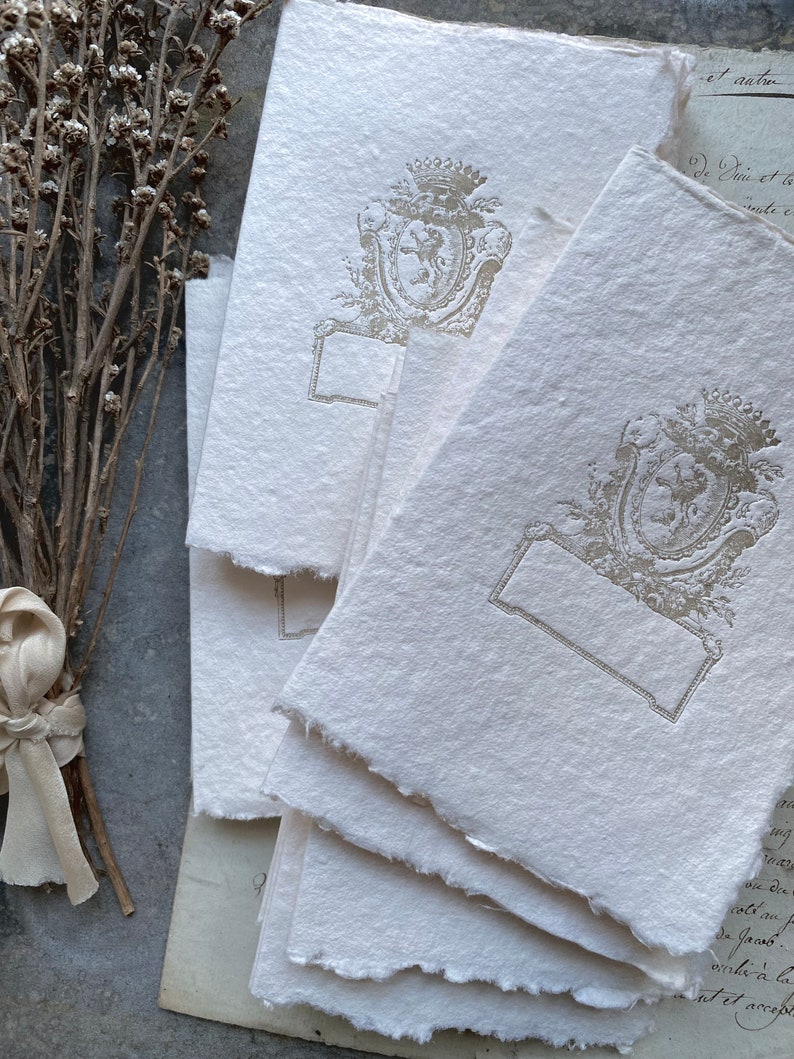 Puede incluir: Una pila de tarjetas de papel en blanco, de color blanquecino, con textura, con un escudo decorativo en relieve plateado que presenta una corona y un le&oacute;n. El escudo est&aacute; rodeado de un dise&ntilde;o floral. Las tarjetas tienen un borde irregular y desgarrado.