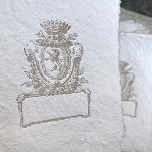 Puede incluir: Una tarjeta de papel blanco con un dise&ntilde;o en relieve marr&oacute;n de un le&oacute;n en un escudo con una corona encima. El escudo est&aacute; rodeado de hojas y flores. Hay un espacio rectangular en blanco debajo del escudo.