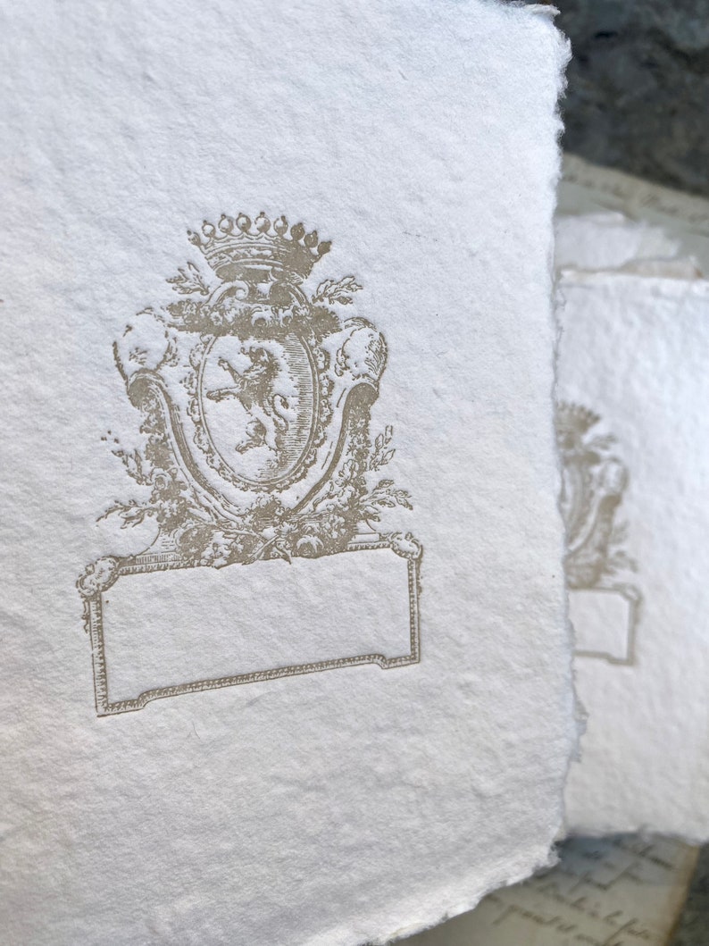 Puede incluir: Una tarjeta de papel blanco con un dise&ntilde;o en relieve marr&oacute;n de un le&oacute;n en un escudo con una corona encima. El escudo est&aacute; rodeado de hojas y flores. Hay un espacio rectangular en blanco debajo del escudo.
