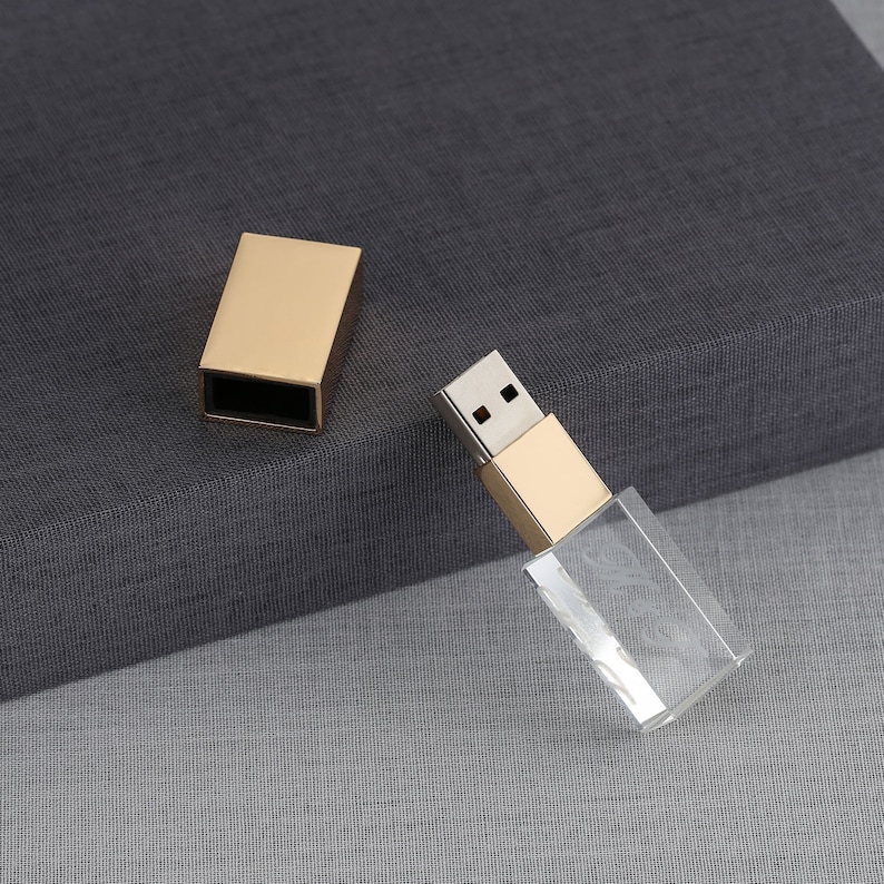 May include: A gold USB drive with a clear crystal casing. The drive is on a gray fabric surface.
