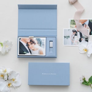 May include: A light blue wedding photo box with a magnetic closure, containing wedding photos and a USB drive. The box is open, revealing a photo of a couple. A closed box with the text "Rebecca & Davin" is also present.