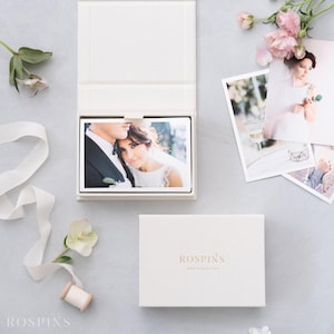 May include: A white rectangular presentation box with the gold text ROSPINS printed on the lid. The open box contains a photograph of a couple, while two additional loose photographs of a person in a white dress are displayed nearby.