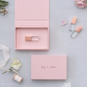 Può includere: Una scatola rosa con un'unità USB in oro rosa all'interno. La scatola è aperta e l'unità USB è visibile. La scatola è su una superficie grigio chiaro. C'è una scatola rosa più piccola con il testo "Bryce & Andrew" su di essa. Ci sono anche tre unità USB in oro rosa sulla superficie.