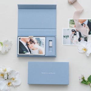 Puede incluir: Una caja de presentación rectangular de color azul claro con tapa abatible, que contiene una fotografía de una pareja y una pequeña unidad USB rectangular. La tapa tiene grabado el texto REBECCA & DAVIN. Hay dos fotos adicionales y flores blancas.