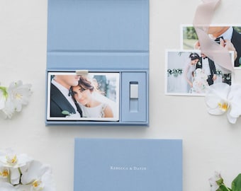 May include: A light blue rectangular presentation box with a hinged lid, containing a photograph of a couple and a small rectangular USB drive. The box cover is embossed with the text REBECCA & DAVIN. Two additional photographs and white flowers are nearby.