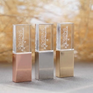 Puede incluir: Tres memorias USB rectangulares con tapas de acrílico transparente y bases metálicas en oro rosa, plata y oro. Cada tapa transparente presenta el texto grabado en blanco ROSPINS Handmade junto a un pequeño gráfico de una rama.