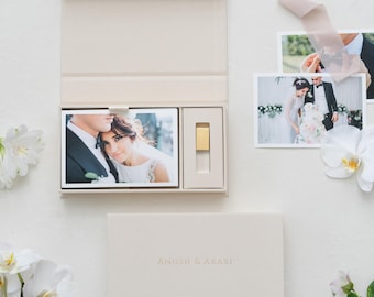 May include: A beige presentation box with a magnetic lid, containing a printed photograph of a couple and a gold USB drive. The box lid is embossed with the text ANOSH & ARABI. Two additional loose photographs and white orchid flowers are nearby.
