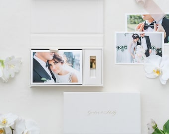 May include: A white rectangular presentation box with a hinged lid, containing a stack of wedding photographs and a gold-toned USB drive. The box lid features the gold embossed text "Gretchen & Shelby" and is surrounded by white orchid flowers.
