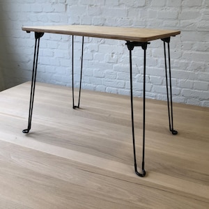 Puede incluir: Una pequeña mesa rectangular de madera con acabado marrón claro. La mesa tiene cuatro patas negras de horquilla. La mesa mide aproximadamente 76 cm de largo y 38 cm de ancho.