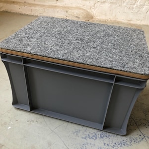 Peut inclure: Une poubelle de rangement en plastique gris avec un dessus en bois recouvert de tissu gris. La poubelle est posée sur un sol en béton.