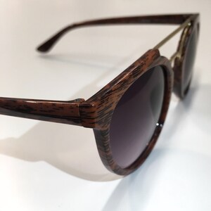 May include: Brown wood-grain sunglasses with a gold metal bar across the top of the frame.
