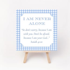 May include: A small, square sign with a light blue gingham border and the text "I AM NEVER ALONE." The sign includes a quote from Isaiah 41:10. It is displayed on a small wooden easel.