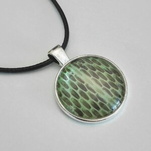 Könnte beinhalten: Eine runde Anhängerhalskette mit silberfarbener Lünette und schwarzem Band. Der Anhänger zeigt ein grün-graues Muster, das Schuppen ähnelt. Die Halskette ist ein schlichtes, aber stilvolles Accessoire.
