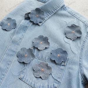 Könnte beinhalten: Ein hellblaues Jeanshemd mit sechs aufgenähten, blütenförmigen Stoffapplikationen mit gewelltem Rand auf der Vorderseite. Jede Blume besteht aus dunklerem Jeansstoff und ist mit einem kleinen, mittigen orangefarbenen Kreuzstich befestigt.