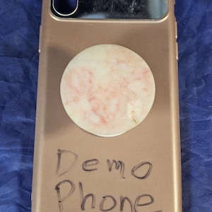 May include: Rose gold phone case with a circular, marble-patterned phone grip. The case has a camera cutout and the words "Demo Phone" are written on the back in black marker.