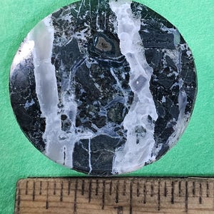 May include: A round, polished stone with a dark green and white marbled pattern. The stone has a smooth surface and is set against a green background. A wooden ruler is in the foreground.