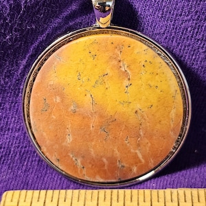 May include: A round pendant with a yellow and orange agate stone set in a silver bezel. The stone has a natural, organic pattern with brown veins and streaks.