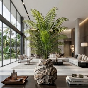 May include: A green palm plant with feathery fronds is potted in a textured, gray, rock-like stone vase. The vase has a rugged, layered appearance with deep crevices and a wide base, sitting on a dark surface alongside a tray and books.