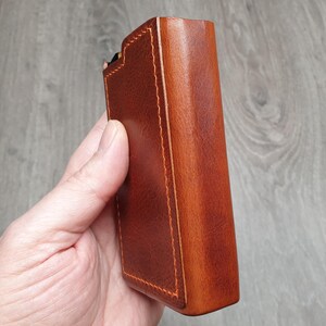 Puede incluir: Una funda rectangular de cuero marr&oacute;n con costuras naranjas. La funda se sostiene en una mano y parece ser una funda protectora para un dispositivo. El cuero tiene un acabado liso y pulido.
