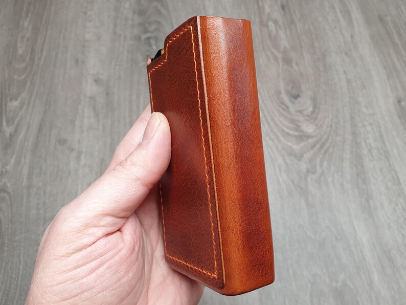 Puede incluir: Una funda rectangular de cuero marr&oacute;n con costuras naranjas. La funda se sostiene en una mano y parece ser una funda protectora para un dispositivo. El cuero tiene un acabado liso y pulido.